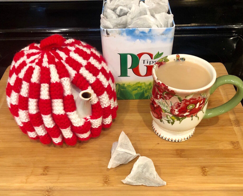 P-G Tips tea in a tin, with 2 loose tea bags, milky tea in a flower cup and a white teapot with a red and white tea cosy on it