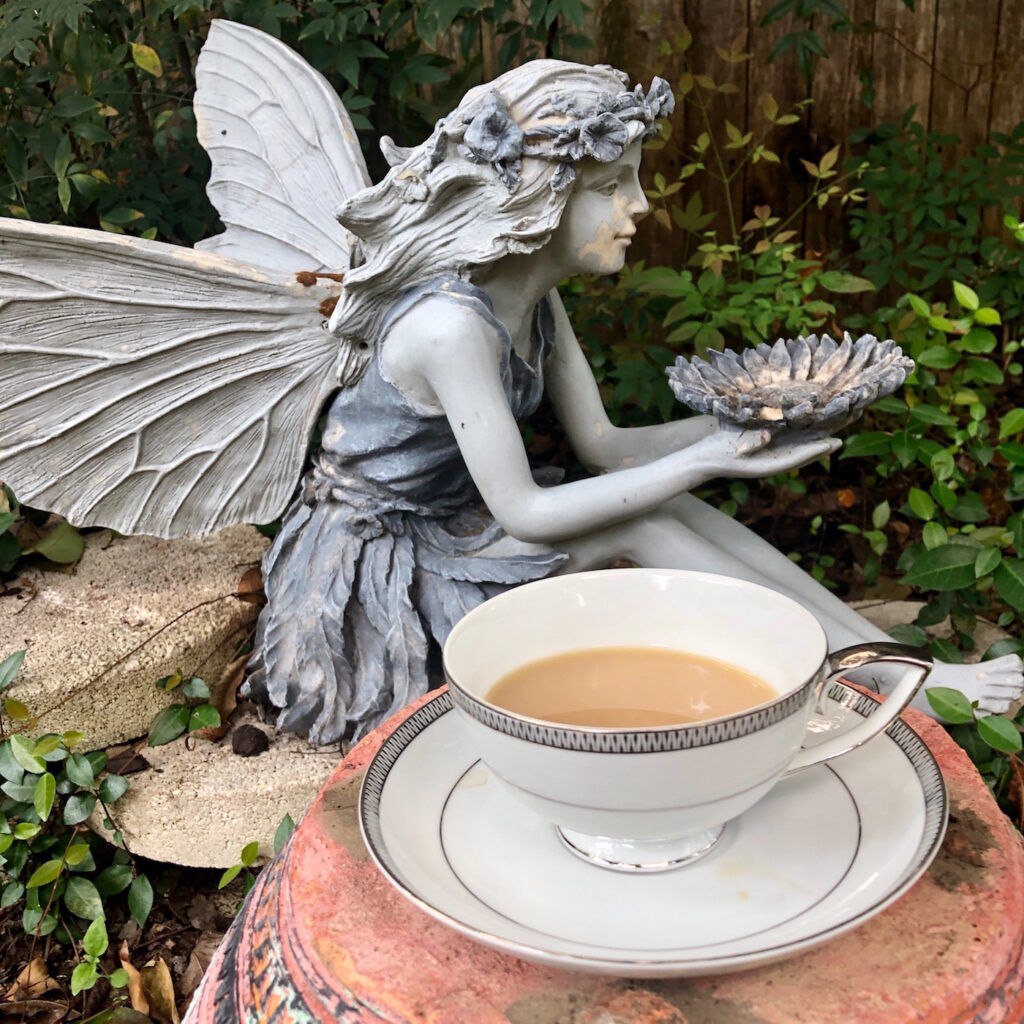 Tea in a white tea cup and saucer with silver rim next to a fairy statue in a garden