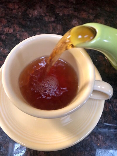 Tea pouring from a green teapot spout into a yellow Fiestaware cup and saucer
